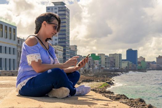 Woman sitting on the Malecon in Havana in front of the sea using a cell phone