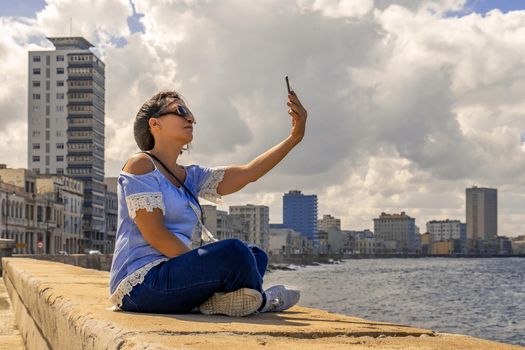 Woman sitting on the Malecon in Havana in front of the sea using a cell phone