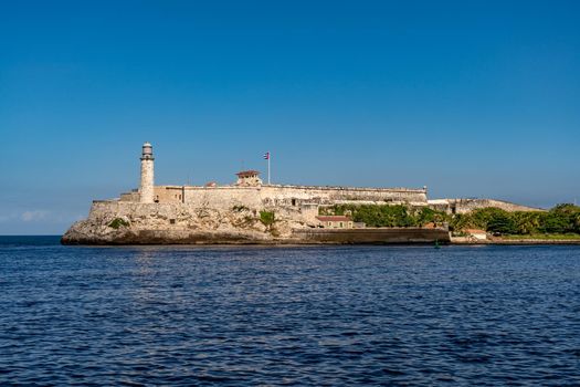 Havana Cuba. November 25, 2020: Horizontal photo of the Morro de la Habana fortress and the lighthouse surrounded by the sea at the entrance to the bay of Havana. In the center the Cuban flag waving on a pole. Famous and tourist place