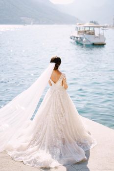 The bride in a cream dress stands on a pier near the sea and admires the mountains and moored boats. The plume of the dress is laid out on the floor and a long veil develops in the wind.