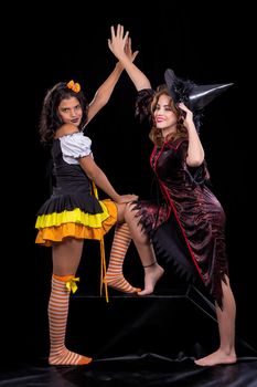 girl dressed as doll and another girl dressed as a witch both portrayed on black background