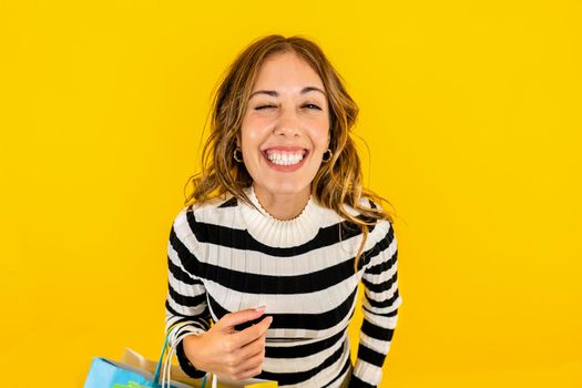 Young beautiful Caucasian woman laughs hard and winking as she looks at the camera happy with her online purchases. Studio shot of happy model holding shopping bags on yellow background for copy space