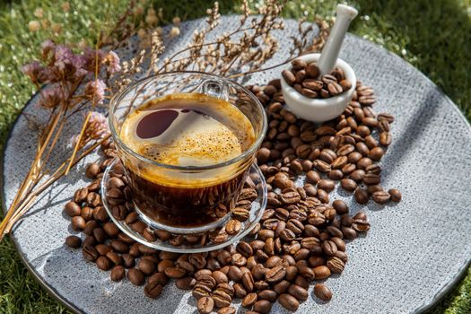 Coffee style, Black coffee in glass cup with coffee beans roating and dried flower on Ceramic plate at lawn in the morning.