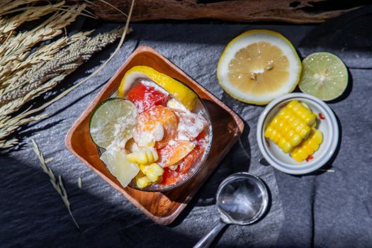 Tropical fresh fruit salad with milk and oatmeal in a glass cup served with corn on dark background. Sweet healthy breakfast, Healthy vegan meal. Top view, Selective focus.