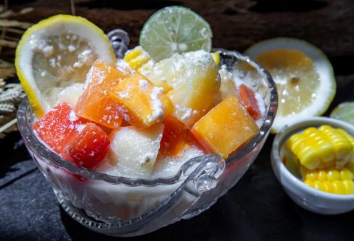 Tropical fresh fruit salad with milk and oatmeal in a glass bowl served with corn on dark background. Sweet healthy breakfast, Healthy vegan meal. Selective focus.