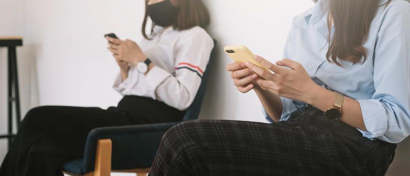 Two Asian young woman chatting in smartphone and wearing mask sitting distance protect from COVID-19 viruses for social distancing for infection risk