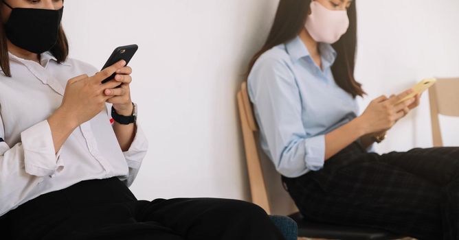 Two Asian young woman chatting in smartphone and wearing mask sitting distance protect from COVID-19 viruses for social distancing for infection risk