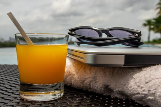 Relaxing moment by the pool. Take a break from working on laptop and enjoy the fresh orange juice for a refreshment.  Outdoor swimming pool background. leisure activity.