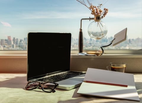 Working space with laptop, notebook, pencil, sunglasses, coffee cup, dried flower vase by the window with city views. Remote working concept or Business workspace at home or office.