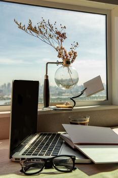 Working space with laptop, notebook, pencil, sunglasses, coffee cup, dried flower vase by the window with city views. Remote working concept or Business workspace at home or office.