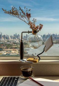 Working space with laptop, notebook, pencil, sunglasses, coffee cup, dried flower vase by the window with city views. Remote working concept or Business workspace at home or office.