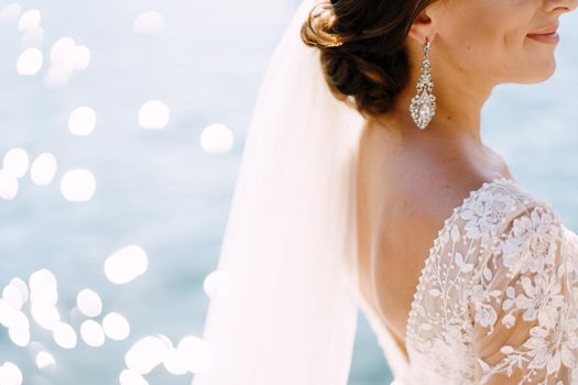 Close-up of the bride's neck and neck, half the profile of the face. Beautiful large earring in the ears, shoulder with white lace dress against the background of the glare of water.