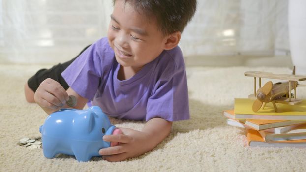Happy Asian kid boy preschool putting pin money coins into blue faced piglet slot. Little child putting coin into piggy bank for saving with pile of coins at home, Investment education concept