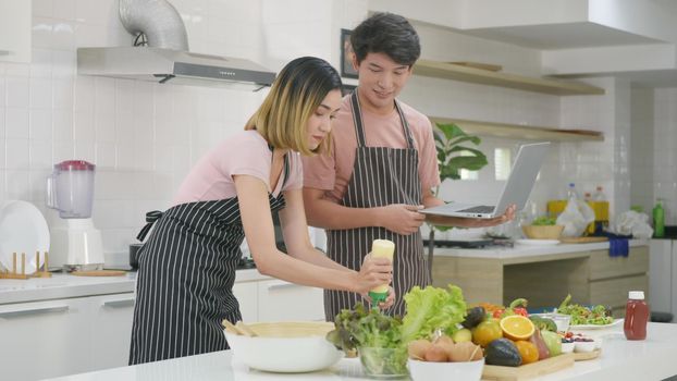 Happy Asian beautiful young family couple husband and wife cooking food vegetable salad in kitchen together at home. The man and woman according online cooking class to recipe on laptop computer