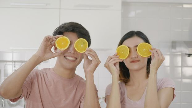 Happy Asian beautiful young family couple husband and wife enjoying smile and laugh holds a cut orange in front of the guy's eyes spending time together in kitchen at home.