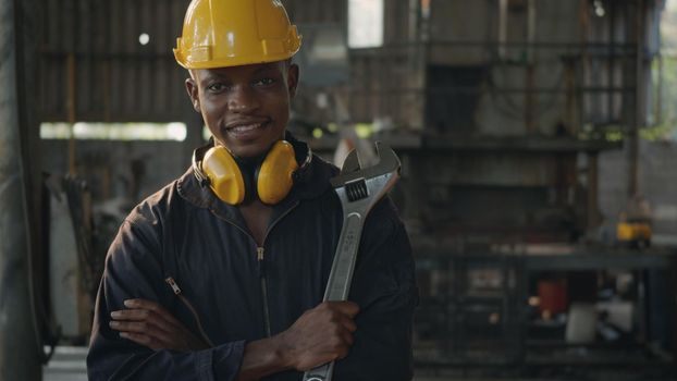 Portrait American industrial black young worker man smiling with yellow helmet in front machine, Engineer standing holding wrench tools and arms crossed at work in the industry factory.