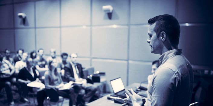 Speaker giving a talk on corporate business conference. Unrecognizable people in audience at conference hall. Business and Entrepreneurship event. Blue toned black and white.