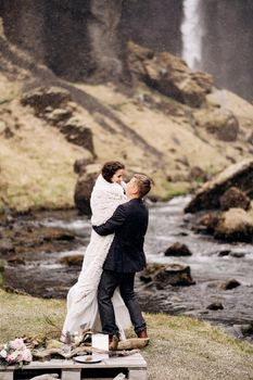 Wedding couple on the banks of a mountain river. The groom wears the bride in a woollen plaid in his arms.