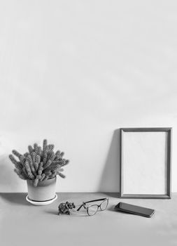 On a table against a white wall is a wooden photo frame and a potted cactus flower. Next to it are glasses and a smartphone. Front view, copy space