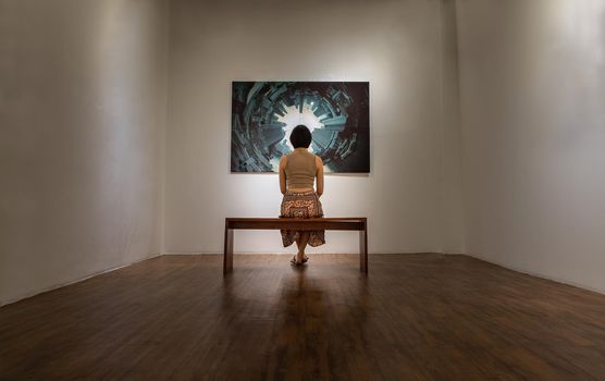 Bangkok, Thailand - 24 Feb 2021 : Back view of young asian woman sitting on bench is interested in photograph displayed on white wall at art gallery exhibition hall. Concept of fine art in photographic gallery. Selective focus.