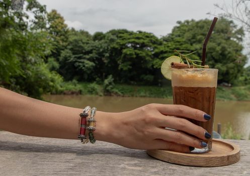 Female hand holding Iced Honey lemon coffee. A glass of coffee mixed with honey and lemon with river view. Refreshing summer drink concept. Selective focus.