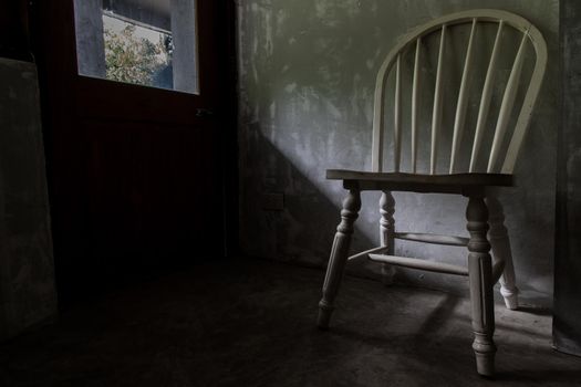 A white wooden chair standing in an empty room under the light from the door. Selective focus.