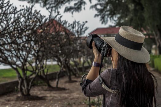 Young woman wearing a white hat and taking a camera to take pictures of the scenery on the sea beach. Traveling and photography. Focus and blur.
