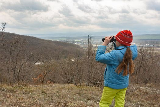 A girl examines a mountain landscape through binoculars, view from the back