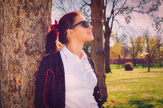 Beautiful casual smiling young girl with sunglasses leaning against a tree in the park, enjoying the sunshine. Young woman leaning against a tree in a city park on a sunny spring day. Nature concept.