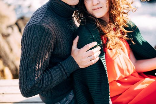 The guy in the knitted sweater, tightly hugs the girl in a red dress and a green sweater by the shoulders.