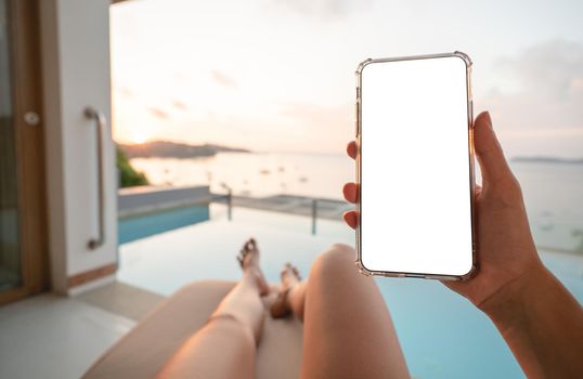 Woman holding mobile phone with blank screen while laying on sunbathing bed with beach view.