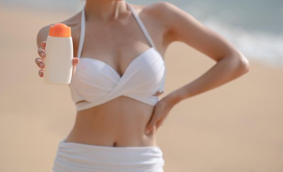 Woman in white bikini holding sunscreen bottle in hand on the beach.