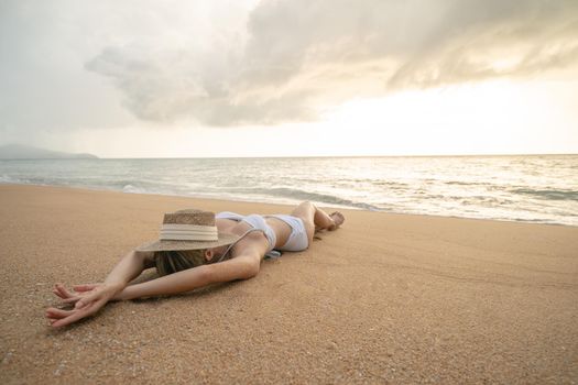 Woman in bikini lying down on sand beach with straw hat cover her face, relaxing sunbathing.