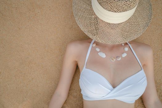 Woman in white bikini lying on sand beach making necklace from sea shell, straw hat covering her face.