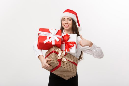 Business Concept - Beautiful young caucasian business woman with santa hat holding a lot of gift boxes and showing thump up.