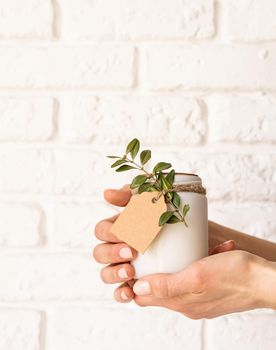 Home decoration and interior. Female hand holding beautiful white candle with fresh leaves on white brick wall background