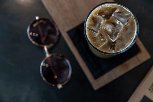 Cold coffee in a tall glass with ice cubes on wooden background. Cold summer drink on a black background with copy space. Focus and blur.