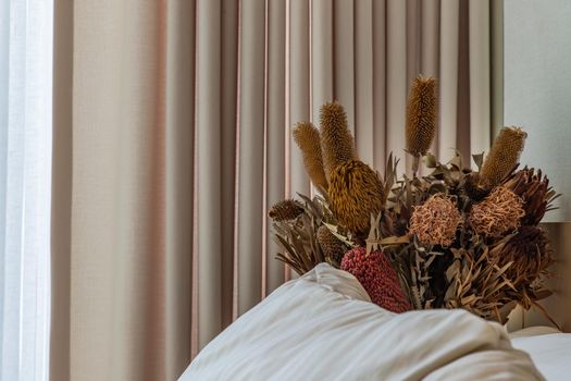 Dry flowers on simple nightstand table with the mattress covered white bed in bedroom. Selective focus.