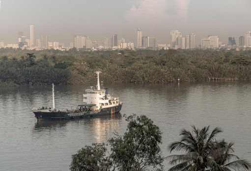 Bangkok, thailand - Nov 20, 2020 : A cargo ship parked in the middle of the river and in front of a green forest with skyscrapers at evening by the Chao Phraya River. Selective focus.