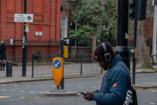 London, United Kingdom - APR 05, 2019 : A young black english man wearing black headphone and listening to music on his smart phone while waiting for the green light to cross the street of London.