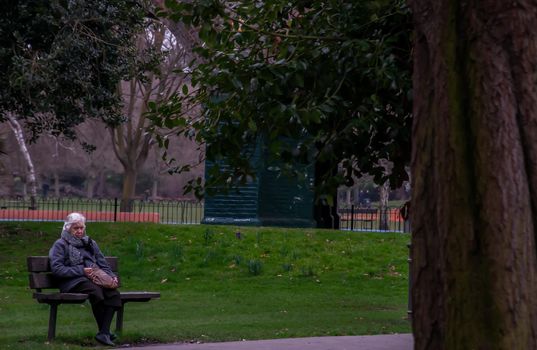 London, United Kingdom - APR 12, 2019 : Elderly caucasian woman sitting on wooden chair in park bench. No focus, specifically.