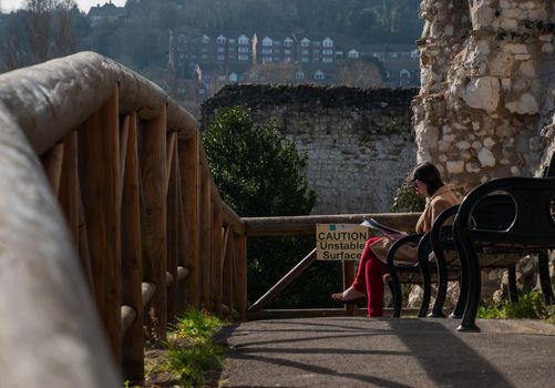 Guildford, United Kingdom - May 05, 2019 : A young woman sitting reading book on wooden bench in Guildford Castle and Grounds. No focus, specifically.