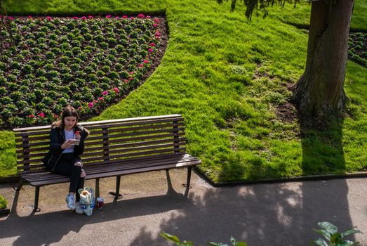 Guildford, United Kingdom - May 05, 2019 : A young woman sitting reading text message on cell telephone on wooden bench in green park. No focus, specifically.