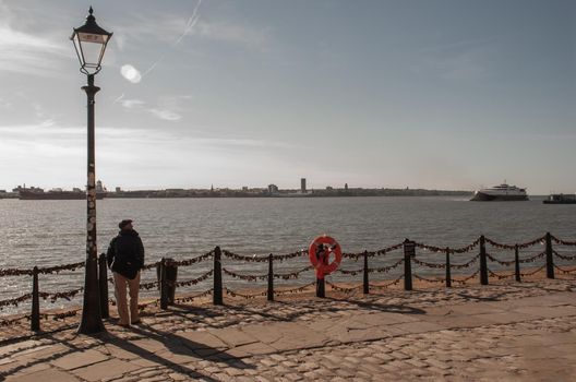 Liverpool, United Kingdom - 03 Jan 2019 : The back of Lonely young man standing under the Street lamp on the river walk and looking at skyline. No focus, specifically.