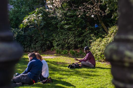 London, United Kingdom - APR 21, 2019 : Young people sit and relax on a green lawn in a park. Summer afternoon in the park. No focus, specifically.