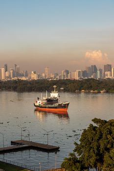 Bangkok, thailand - Nov 20, 2020 : A cargo ship parked in the middle of the river and in front of a green forest with skyscrapers at evening by the Chao Phraya River. Selective focus.