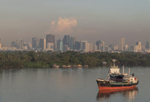 Bangkok, thailand - Nov 20, 2020 : A cargo ship parked in the middle of the river and in front of a green forest with skyscrapers at evening by the Chao Phraya River. Selective focus.