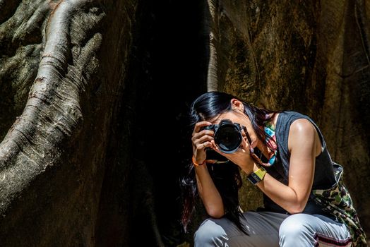 Young woman with Shoulder bag and using a camera to take photo Giant big tree, Size comparison between human and giant big tree in Ban Sanam of Uthai Thani Province, Thailand, nature background.