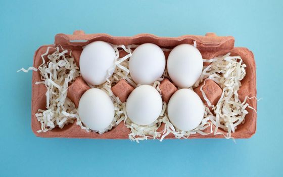 Six white fresh organic eggs in a beige eco-friendly cardboard egg container on a blue background. Easter concept, ecology, zero waste.