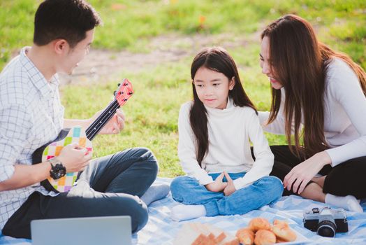 Happy Asian young family father, mother and children having fun and enjoying outdoor together sitting on the grass party with playing Ukulele during a picnic in the garden park on a sunny day